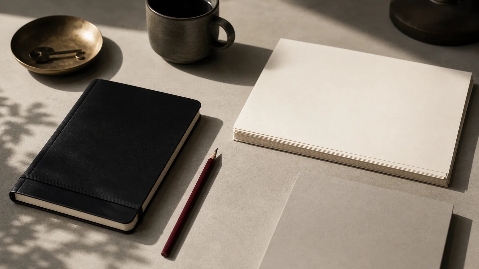 A clean desk with blank paper, a black notebook, a pencil, and a coffee cup in soft window light.